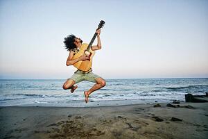 Fotografia Mixed Race man playing guitar and jumping at beach, Peathegee Inc, (40 x 26.7 cm)